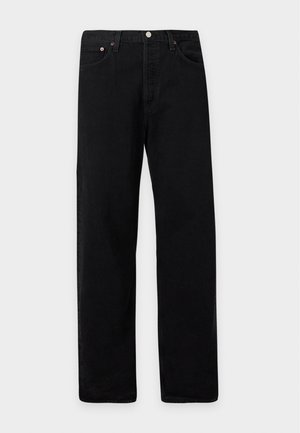 Jeune homme avec une chemise noire à manches longues superposées, un pantalon noir et des chaussures noires, debout devant un fond clair uni.