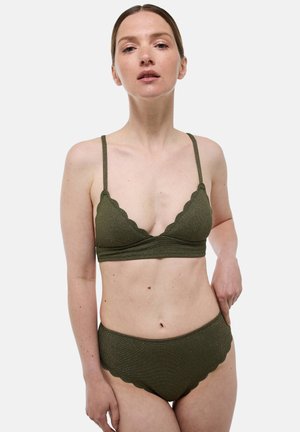 Femme à la peau claire et aux cheveux bruns portant un bikini vert texturé avec des bords festonnés, se tenant devant un fond blanc uni.