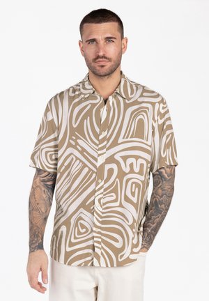 Mężczyzna z krótkimi włosami i tatuażami na obu ramionach, ubrany w beżową i białą koszulę na krótki rękaw z abstrakcyjnym wzorem i guzikami.