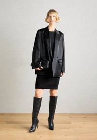 Femme portant un blazer noir surdimensionné, des bottes noires montantes jusqu'aux genoux, une robe noire ajustée, tenant un petit sac à main noir, debout sur un sol en bois.