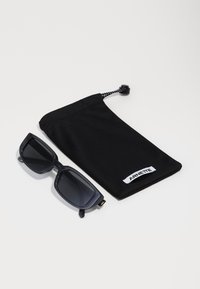 Lunettes de soleil rectangulaires noires avec des verres sombres à côté d'une pochette en tissu noir avec un cordon de serrage et une étiquette de logo blanche.