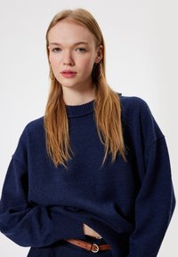 Granatowy, oversize sweter z prążkowanym okrągłym dekoltem i fakturowanym splotem. Modelka ma na sobie brązowy pasek, a widać także długie, proste włosy.