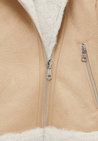 Camelkleurig suède jack met een voering en kraag van nepbont. Voorzien van dubbele zilveren ritsen en een zachte, gestructureerde afwerking aan de zoom.