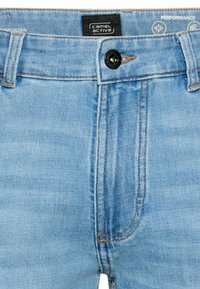 Lyseblå denimjeans med sort knap, tan-syninger, bæltestropper og et Camel Active mærke på linningen.