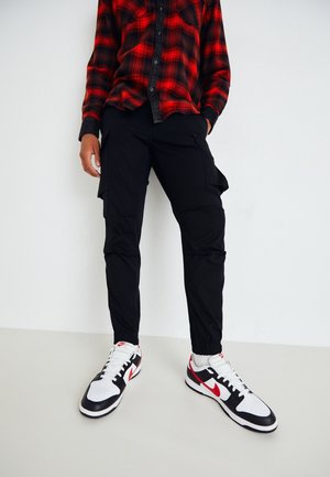 Persoon draagt zwarte cargobroek, witte sneakers met rode Nike-swoosh en een rood-zwarte geruite blouse tegen een effen witte achtergrond.