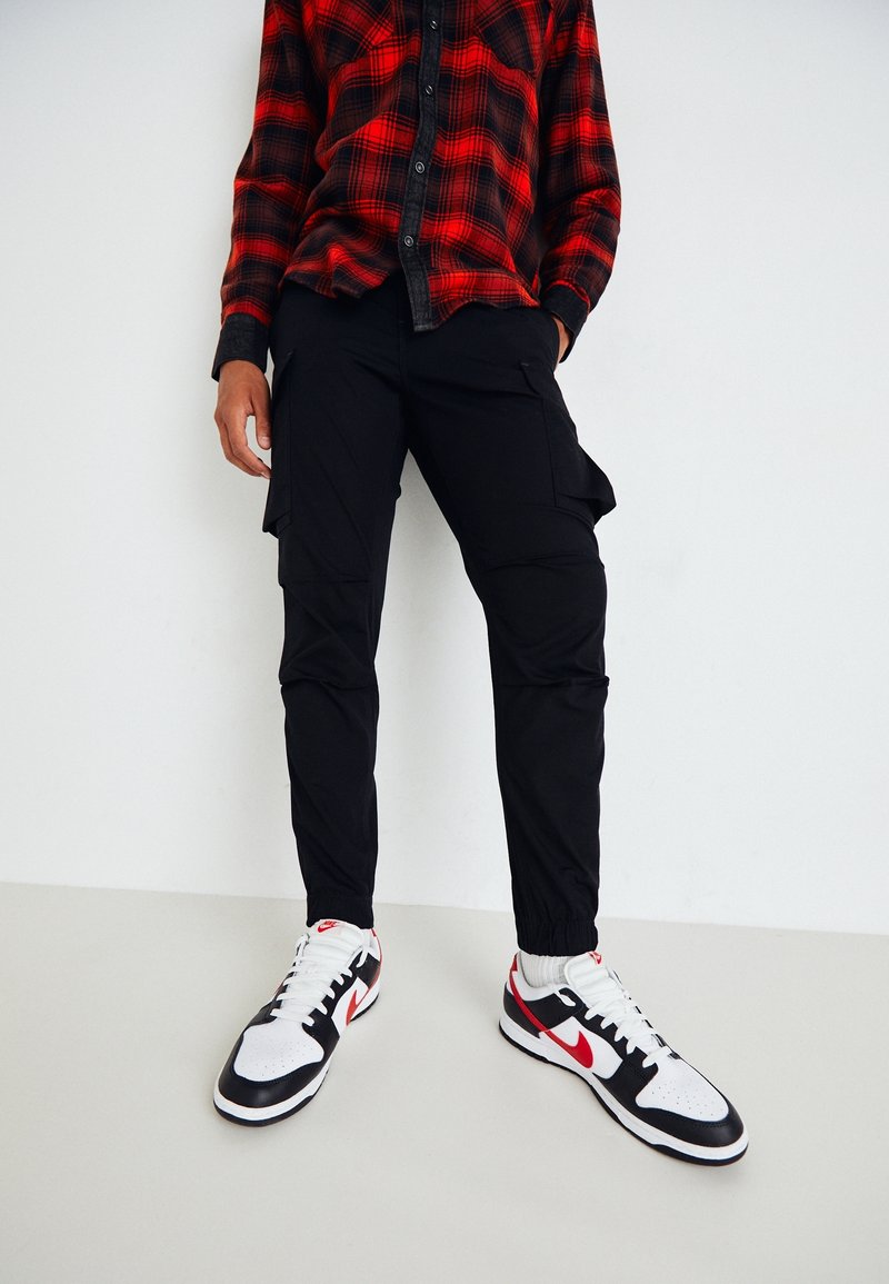 Personne portant un pantalon cargo noir, des baskets blanches avec le swoosh Nike rouge, et une chemise à carreaux rouge et noir sur un fond blanc uni.