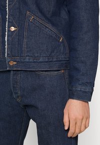 Tmavě modrá džínová bunda s kontrastním prošíváním, měděným kováním a přední kapsou. Bunda má strukturovaný design a texturovanou tkaninu.