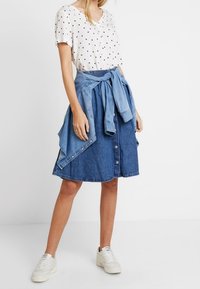 Top corto de manga corta blanca con un patrón de puntos negros, combinado con una falda de mezclilla azul con botones y una chaqueta de mezclilla atada. Zapatillas blancas completan el atuendo.