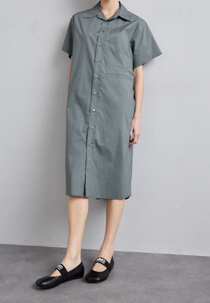 Personne portant une robe grise boutonnée à manches courtes et longueur genoux, associée à des ballerines Mary Jane noires avec des brides blanches avec logo.