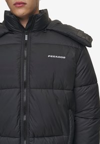 Svart puffjacka med hög krage, med dragkedja fram, synlig vaddering och varumärket "PEGADOR" tryckt i vitt.