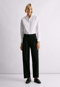 Camicia bianca con bottoni, pantaloni neri plissettati con una trama maculata e mocassini neri. Taglio semplice e su misura con linee pulite.