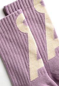 Een paar geribbelde lavendelkleurige sokken met een groot crèmekleurig sleutelontwerp gebreid aan de zijkanten, plat gelegd op een witte ondergrond.