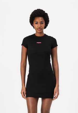 Robe noire ajustée avec des manches courtes, arborant un petit logo rose sur la poitrine. Texture lisse, longueur au-dessus du genou, design minimaliste.
