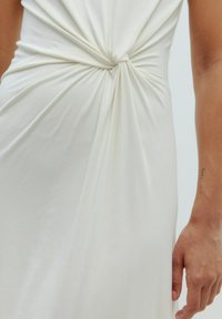 Witte jurk gemaakt van zachte stof, met een gedraaide knoopdetail bij de taille en een soepele drapering die de lichaamsvorm accentueert.