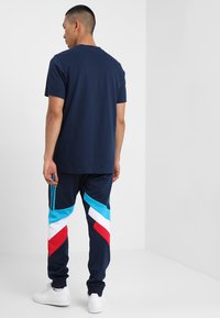T-shirt navy con colletto tondo, abbinato a pantaloni da jogger blu, bianchi e rossi con motivo a blocchi di colore diagonali e tre strisce sui lati.