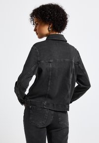 Musta farkkutakki, jossa on klassinen kaulus, pitkät hihat ja näkyvät ompeleet. Takakuvassa näkyy rakenteellinen muoto ja kapea leikkaus.