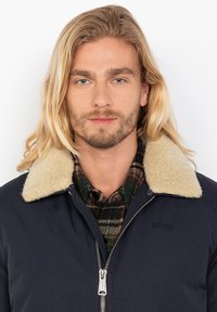 Chaqueta azul marino con un suave cuello de piel beis, cierre de cremallera y logo de la marca. Llevada sobre una camisa de cuadros verde y marrón. Tejido texturizado.