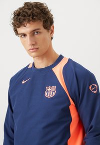 Mørkeblå sweatshirt med lyse orange detaljer, der viser et Nike-logo og FC Barcelona-våbenskjold. Glat stof, rund hals og lange ærmer.