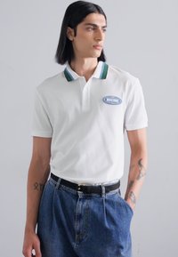 Valge polo särk, millel on roheline ja sinine triibuline krae, rinnal sinine Moschino logo patch. Kantud kõrge vöökohtadega teksashortsidega.