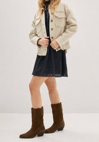 Beige denimjakke med frynset kanter, to brystlommer, båret over en sort kjole med øjelet blonder, parret med brune ruskindsankelstøvler.