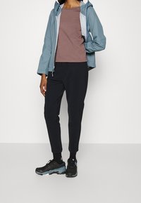 Ljusblå vattentät jacka, brun kortärmad topp, svarta avsmalnande byxor och svarta vandringsskor med turkosa detaljer. Avslappnad utomhusklädsel.