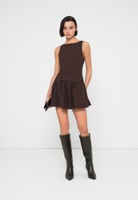 Καφέ αμάνικο φόρεμα με εφαρμοστό μπούστο και φουσκωτή φούστα με πτυχώσεις, με κορδονάκια στη μέση. Φορεμένο με μαύρες μπότες μέχρι το γόνατο.