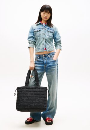 Nuori nainen vaaleassa farkkutakissa ja leveälahkeisissa farkuissa, joka pitää suurta mustaa tikattua laukkua ja punaisia sandaaleja valkoisella taustalla.