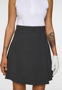 Gonna da golf grigia con pieghe, realizzata in tessuto elasticizzato, abbinata a una maglietta bianca senza maniche. Una mano indossa un guanto da golf bianco.