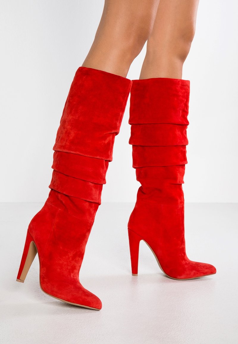 Femme portant des bottes à talons hauts en suède rouge vif avec un design ample et empilé, debout sur un sol blanc devant un fond blanc uni.