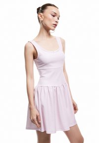 Joven mujer que lleva un vestido sin mangas de cuadros rosas con un borde de encaje, de pie con la cabeza girada ligeramente hacia un lado sobre un fondo blanco.