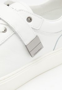 Weißer Ledersneaker mit silbernem rechteckigem Akzent und "Calvin Klein"-Markenlogo, flachen Schnürsenkeln und einer Gummisohle mit subtiler Struktur.
