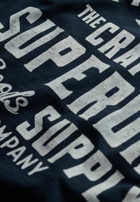 Nærbillede af marineblåt stof med store, slidte hvide bogstaver, der læser "THE CRANE SURF AND SUPPLY COMPANY" i fed, vintage stil skrifttype.