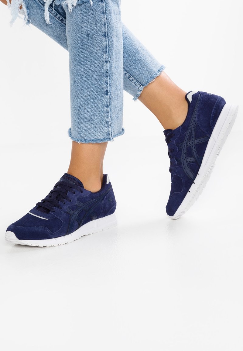 Personne portant un jean bleu clair déchiré et des baskets bleu marine avec des semelles blanches, levant légèrement un pied du sol.