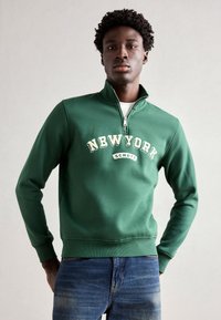 Sweatshirt vert à col zippé avec le texte "NEW YORK" en crème, accompagné de poignets et d'un ourlet côtelés, porté sur un jean en denim.