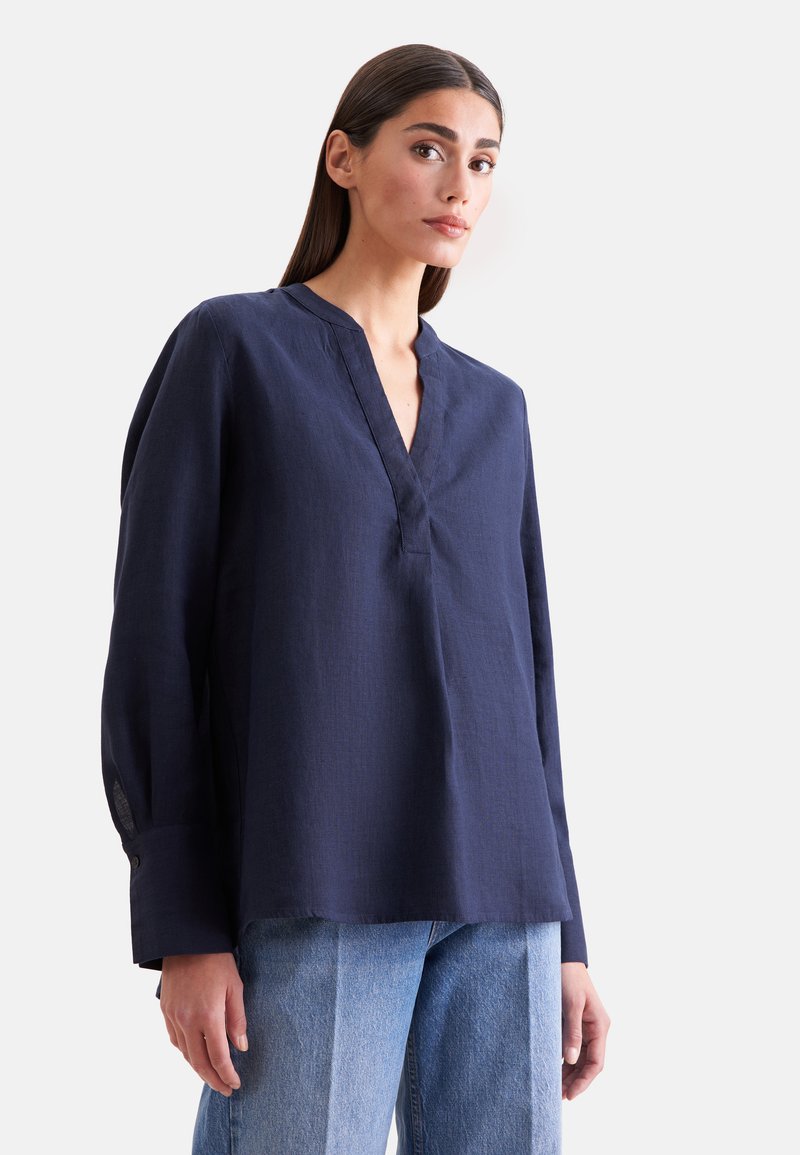 Femme aux longs cheveux foncés portant un chemisier ample bleu marine à col en V et un jean bleu clair sur un fond uni.