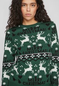 Pull en tricot vert avec des rennes blancs, des flocons de neige et le texte "JOYEUX NOËL !" en noir. Il a un col rond et une bande horizontale à motif.