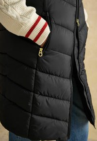 Czarna puchowa kamizelka z zapinanymi kieszeniami, z przeszyciami i złotymi detalami. Kremowy sweter z czerwonymi paskami widocznymi pod spodem.