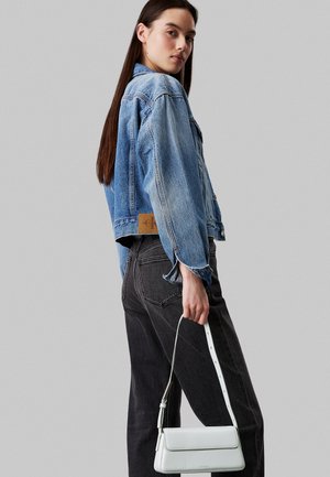 Chaqueta de mezclilla azul claro con un corte corto, combinada con jeans gris oscuro. Un pequeño bolso blanco es sostenido por el modelo.