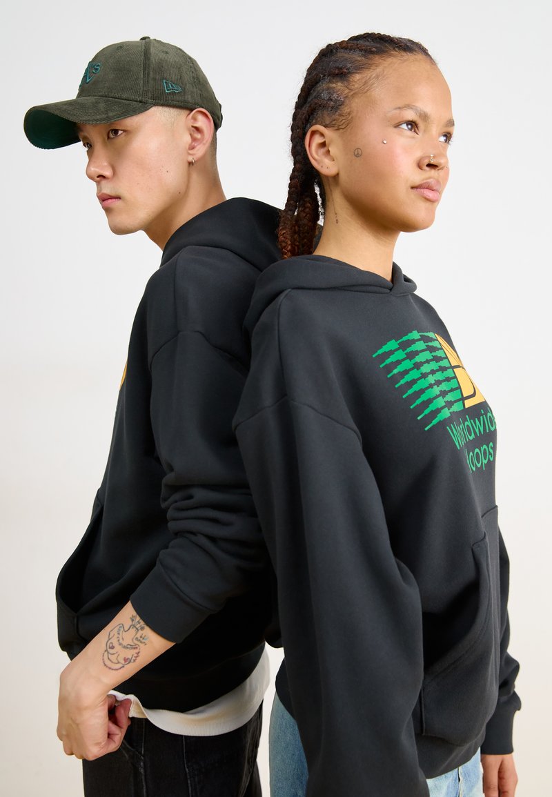 Czarne bluzy z kapturem z zielonymi i żółtymi grafikami, w połączeniu z oliwkową czapką. Oba elementy mają swobodny krój, eksponując tekstury denimu i bawełny.