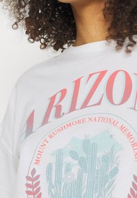 Bílý bavlněný tričko s nápisem "ARIZONA" v červeném a modrém textu, s kruhovou grafikou kaktusů a nápisem "NÁRODNÍ PAMÁTNÍK MOUNT RUSHMORE" níže.