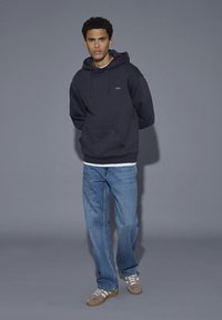 Ung mand iført en sort hættetrøje, blå jeans og sneakers står mod en grå baggrund med hænderne bag ryggen.