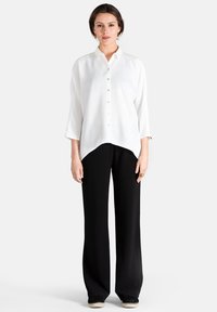 Blusa blanca de botones con cuello y mangas largas, con un dobladillo curvado. Combinada con pantalones de pierna ancha negros y zapatos de esparto.