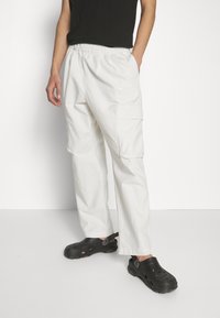 White cargo pants with elastic waistband, side pockets, and subtle Nike "AIR" logo. Worn with black clogs featuring ventilation holes.