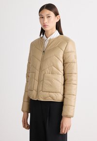 Beige quiltet pufferjakke med et chevron-mønster, lynlås foran og to store påsyede lommer. Glat stof og ribbet krave.