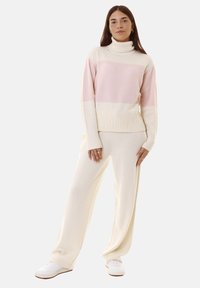 Golfino color crema con collo alto, caratterizzato da strisce orizzontali rosa chiaro; texture morbida, polsini e orlo a costine. Abbinato a pantaloni in bianco sporco coordinati.