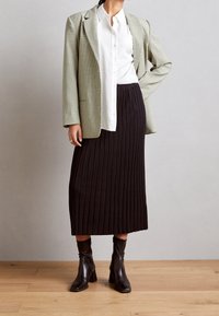 Beige houndstooth blazer, bílá košile s knoflíky, černá plisovaná midi sukně a černé kotníkové boty. Hladké textury, strukturovaný design.