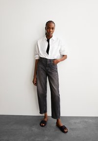 Camicia bianca con bottoni, top nero, jeans scuri a vita alta e morbidi, sandali scivolati scuri. Tessuto liscio con una lieve lucentezza.