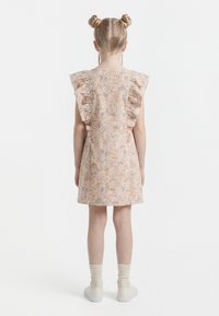 Meisje met dubbele knotjes, gekleed in een knielange bloemenjurk met gerimpelde mouwen, crèmekleurige sokken en witte schoenen, staand met haar rug naar de kijker.