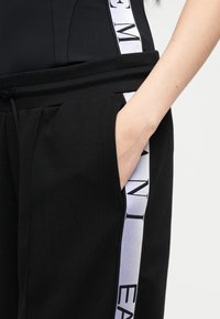 Το χέρι ενός ατόμου ξεκουράζεται στην τσέπη μαύρου παντελονιού με λευκή ρίγα στο πλάι που φέρει μεγάλα μαύρα γράμματα.