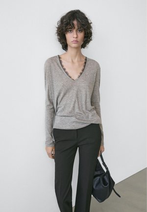 Giovane con capelli ricci che indossa un maglione grigio a scollo a V, pantaloni neri e tiene una borsa nera contro una parete bianca liscia.